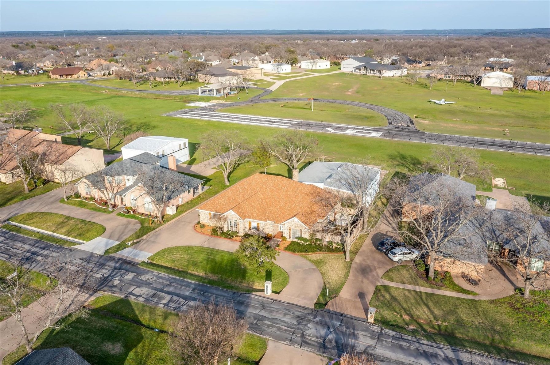 Aerial of a Granbury home in Pecan Plantation with detached outbuildings behind it, a taxiway running out to a paved airstrip, and a light aircraft parked on the grass beyond.