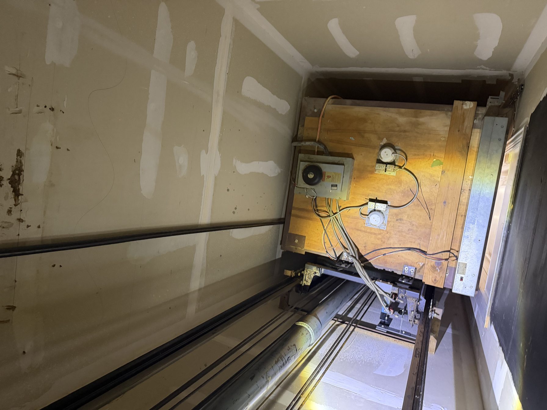 Inside a residential elevator hoistway: drywall on the left, steel guide rails and a hydraulic cylinder running the length of the shaft, and a plywood equipment backboard on the right carrying the disconnect, controller boxes and conduit.