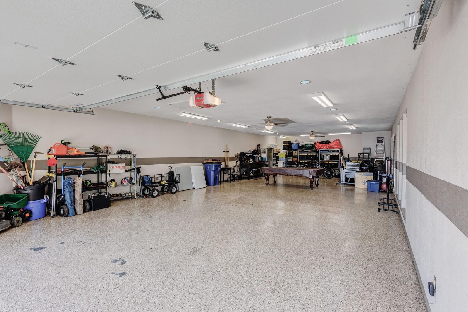 A large finished garage with drywalled and painted walls, a speckled epoxy floor, recessed and fluorescent lighting, ceiling fans, storage shelving along one wall and a billiard table at the far end.
