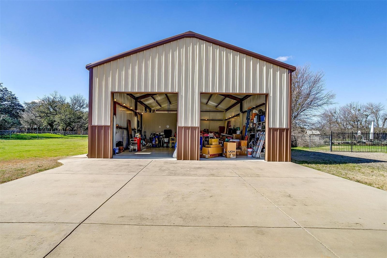 Exterior of a tan metal shop building with two very tall open bay doors, roughly twice the height of an ordinary garage door, opening onto a wide concrete apron.