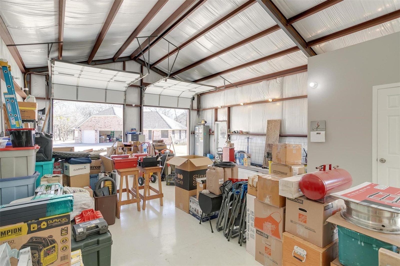 Interior of a metal shop building with two tall roll-up doors standing open, insulated walls, overhead and wall lighting, an electrical panel, a water heater and a door through to a finished room.
