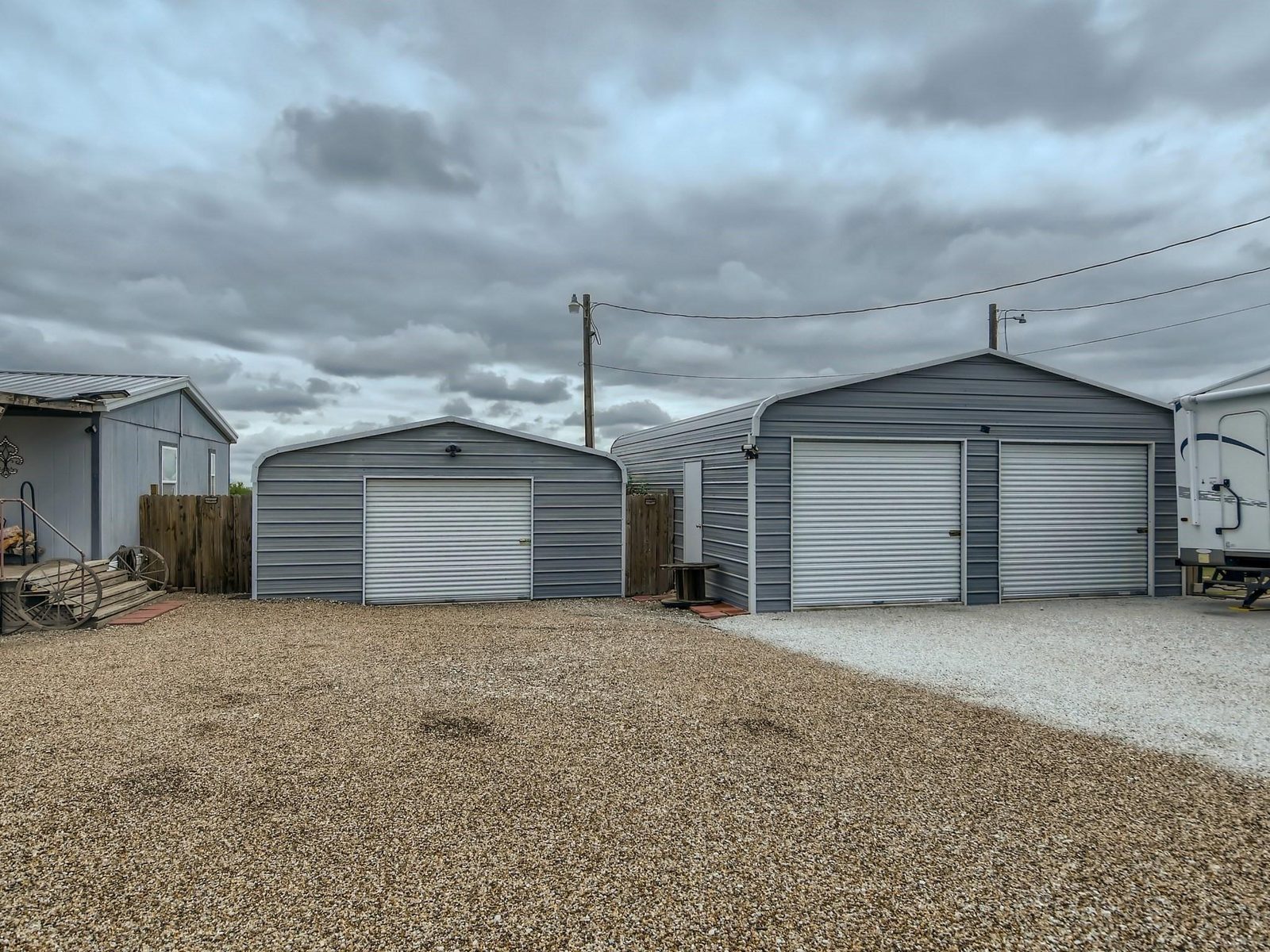 Two prefabricated metal garage buildings with roll-up doors side by side on a gravel yard, a smaller outbuilding to the left and a travel trailer parked at the right.