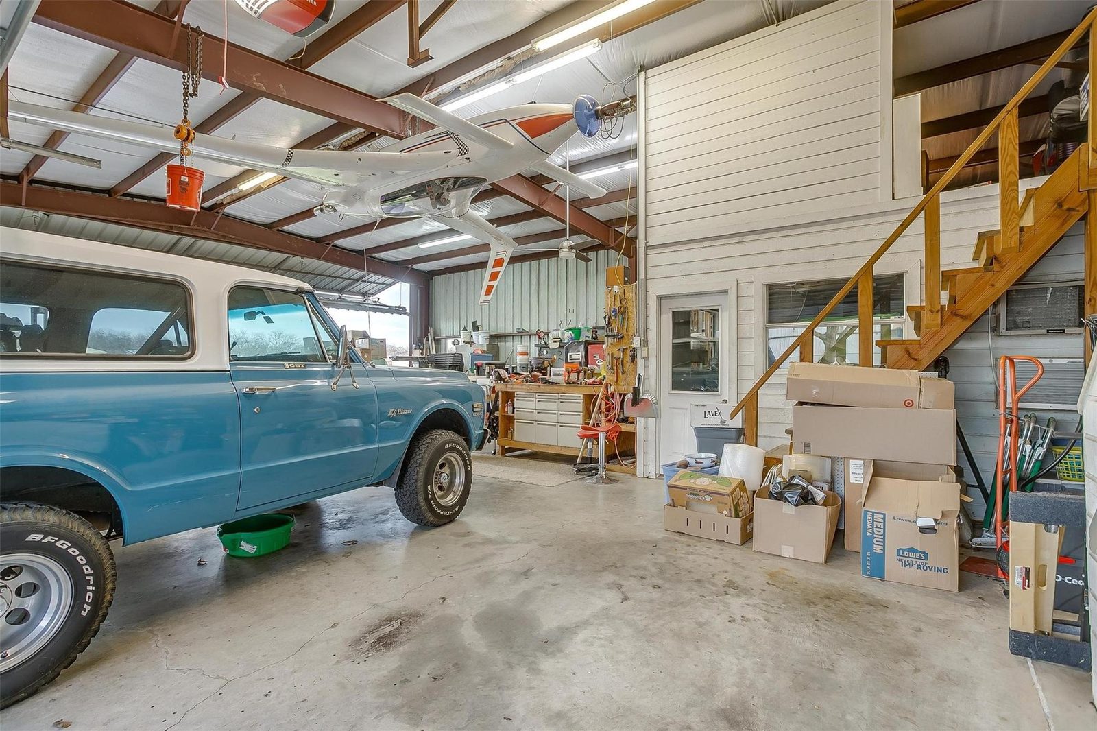 Inside the same shop, a restored vintage four-wheel-drive truck parked under an aircraft suspended from the ceiling, with a workbench and pegboard of tools along the far wall, a chain hoist overhead, and a wooden staircase at the right rising to a second level behind a finished wall.