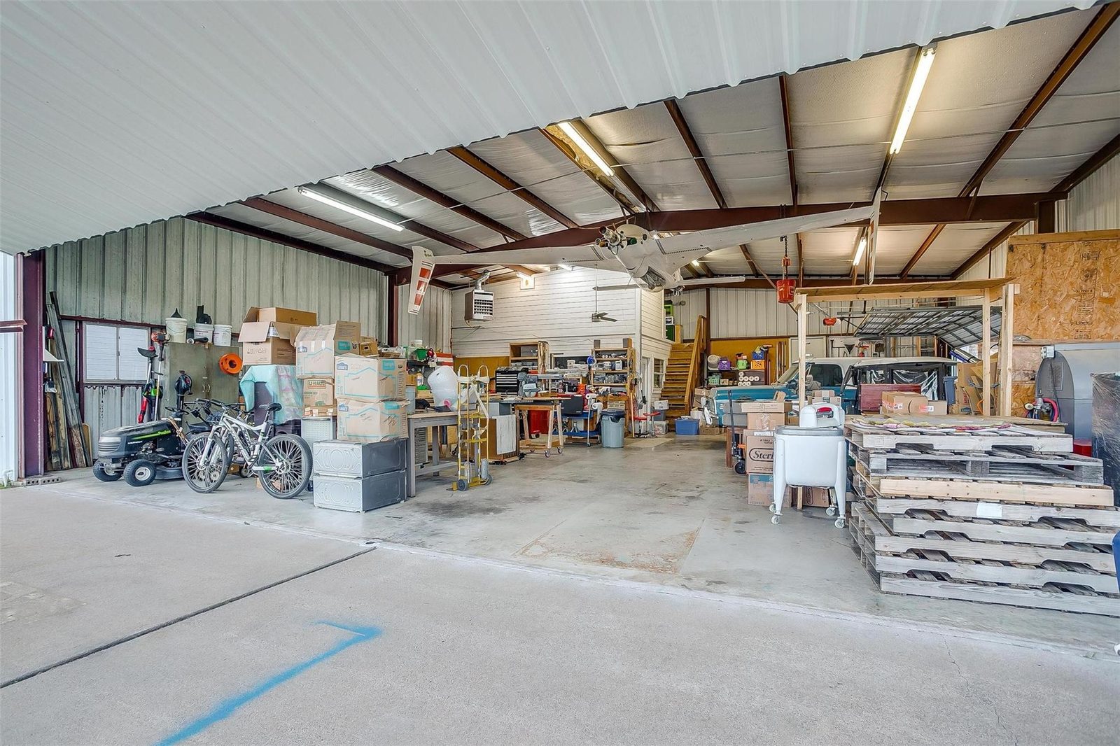 Wide interior view of a large open shop building with an aircraft fuselage and wings suspended from the steel roof structure, a workbench area at the far end, stairs to a second level, a gas unit heater on the wall, and equipment and pallets stored along both sides.